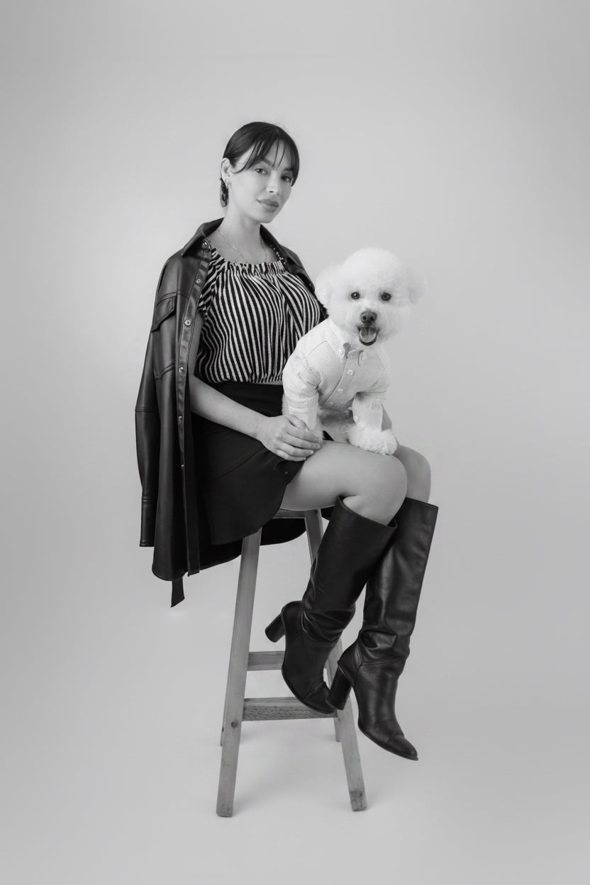 Woman sitting on a stool with a small white dog, wearing a black outfit and knee-high boots.
