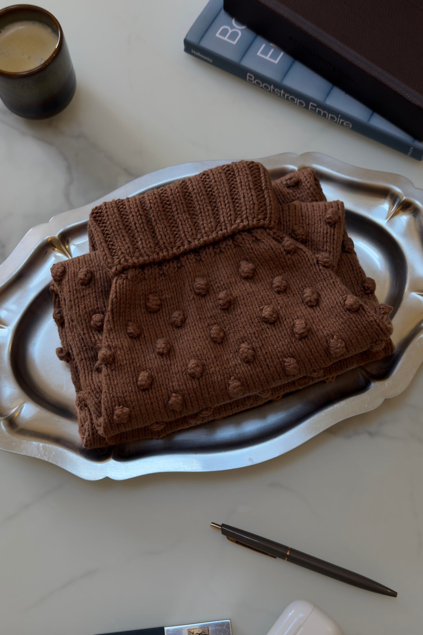 Brown knitted pouch with textured surface on a silver tray, surrounded by books and a candle.