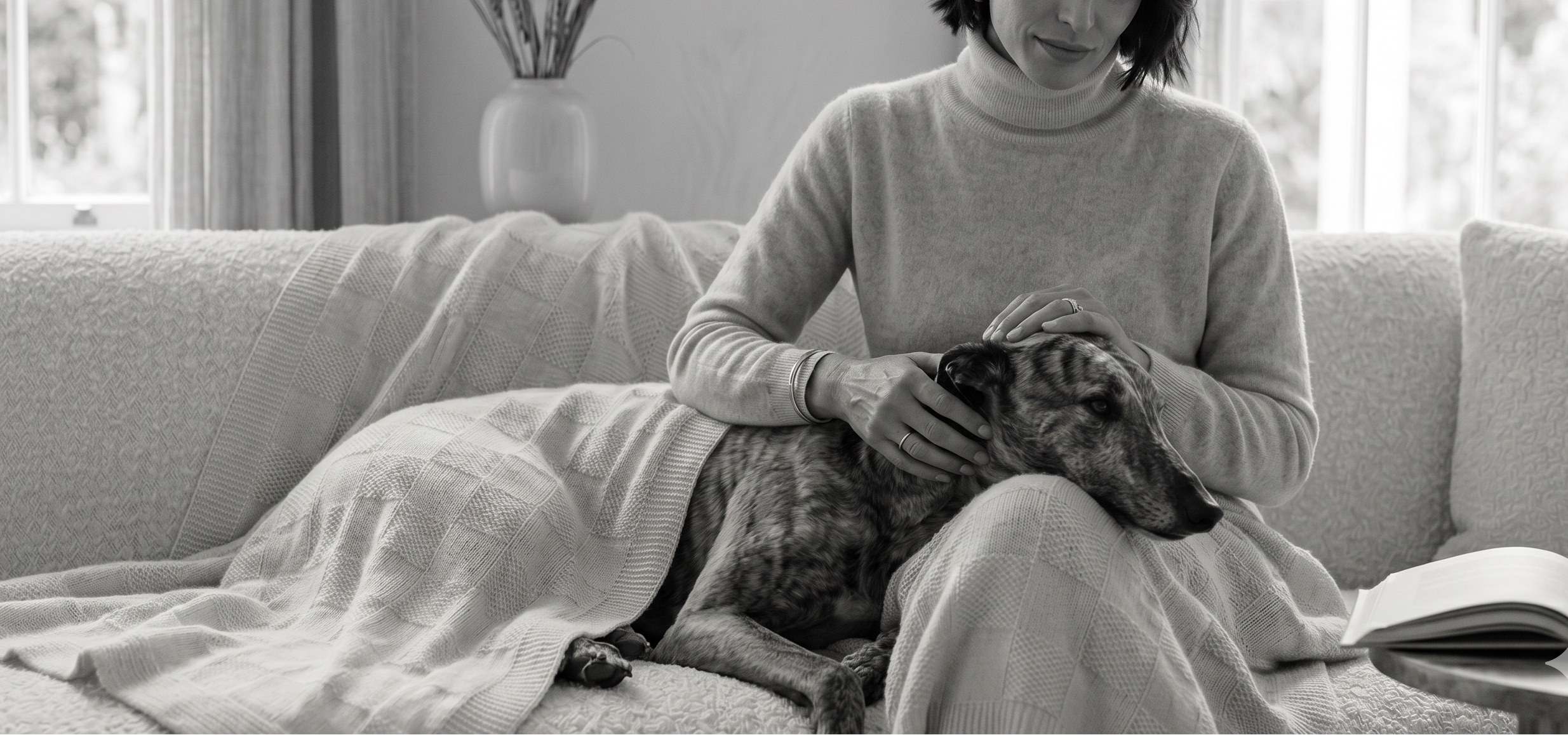 Woman sitting on a couch with a dog, both wrapped in a blanket, in a home setting.